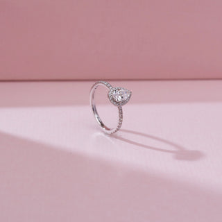 Side angled view of the Pear Halo Radiant Diamond Ring in 14KT white gold, featuring a pear-shaped center diamond encircled by a halo with a slim pavé diamond band on a soft pink background.