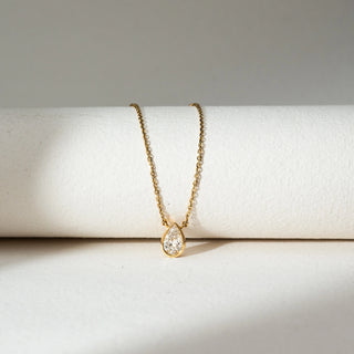 Top view of Pear Bezel Diamond Pendant Necklace in 14KT Yellow Gold, featuring a pear-shaped lab grown diamond in a smooth bezel setting. Placed on a beige backdrop, the design highlights minimal elegance and soft brilliance.
