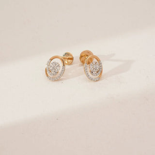 Angled view of yellow gold swirl floral diamond stud earrings with a clustered round diamond centre and pavé-set curved accents, softly lit on a neutral beige surface.