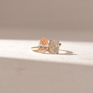 Front view of a diamond ring in 14KT yellow gold, featuring a pear-shaped pink diamond paired with a cushion-cut white diamond, set on a sleek band against a beige backdrop highlighting contrast and brilliance.
