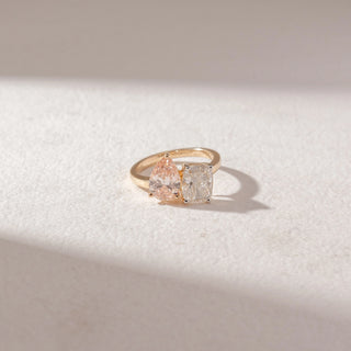 Top view of a diamond ring in 14KT yellow gold, featuring a pear-shaped pink diamond paired with a cushion-cut white diamond, set on a sleek band against a beige surface highlighting contrast and sparkle.
