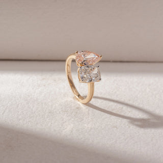 Angled view of a diamond ring in 14KT yellow gold, featuring a pear-shaped pink diamond above a cushion-cut white diamond, set on a sleek band against a beige surface highlighting brilliance and contrast.
