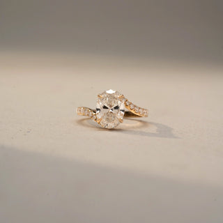 Front view of an oval solitaire engagement ring in 14 KT Yellow Gold, featuring an oval-cut lab-grown diamond in a four-prong setting with a pavé-set diamond band, displayed against a beige backdrop.