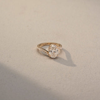 Top view of an oval solitaire engagement ring in 14 KT Yellow Gold, featuring an oval-cut lab-grown diamond in a four-prong setting with a pavé-set diamond band, displayed against a beige backdrop.