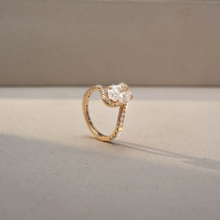 Angled view of an oval halo engagement ring in 14 KT Yellow Gold, featuring an oval-cut lab-grown diamond encircled by a sparkling halo with pavé-set diamonds along the band, displayed against a beige backdrop.