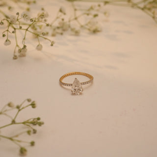 Top view of Reia's Eternal Teardrop Solitaire Diamond Ring in 14KT yellow gold, with a pear-cut lab-grown diamond on a slim pave band, styled with white floral accents.