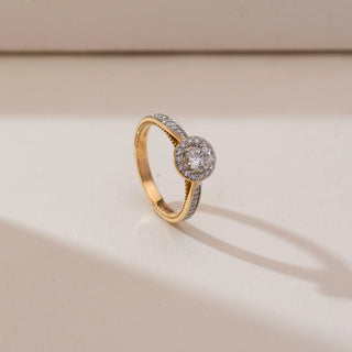 Angled view of Sophisticated Circular Halo Diamond Ring in yellow gold, featuring a round lab-grown diamond encircled by a sparkling halo with diamond-set shoulders, placed on a soft beige backdrop.
