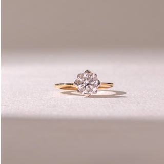 Front view of a diamond ring in 14KT yellow gold, featuring a round center diamond in a floral-inspired prong setting on a sleek band, set on a beige surface highlighting brilliance and timeless elegance.
