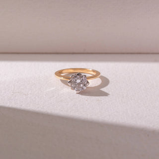 Top view of a diamond ring in 14KT yellow gold, featuring a round center diamond in a floral-inspired prong setting on a sleek band, set on a beige surface highlighting refined sparkle and elegance.

