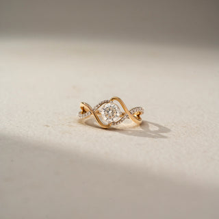 Front view of a swirl solitaire ring in 14 KT Yellow Gold, featuring a round lab-grown diamond framed by flowing pavé-set diamond accents and sculpted crossover gold bands, displayed against a beige backdrop.