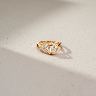 Top view of a swirl solitaire ring in 14 KT Yellow Gold, featuring a round lab-grown diamond framed by flowing pavé-set diamond accents and sculpted crossover gold bands, displayed against a beige backdrop.