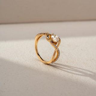 Angled view of a swirl solitaire ring in 14 KT Yellow Gold, featuring a round lab-grown diamond framed by flowing pavé-set diamond accents and sculpted crossover gold bands, displayed against a beige backdrop.