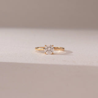 Front view of round diamond solitaire ring in yellow gold with a slim band, set on a soft neutral surface, highlighting classic simplicity and bright, radiant sparkle.
