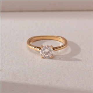 Top view of round diamond solitaire ring in yellow gold with a slim band, set on a soft neutral surface, highlighting classic design and bright, refined sparkle.
