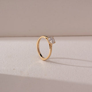 Angled view of round diamond solitaire ring in yellow gold with a slim band, set on a soft neutral background, highlighting elevated setting and classic, radiant sparkle.
