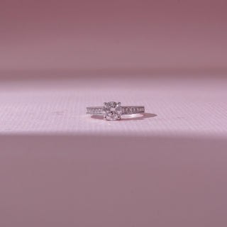 Front view of a round diamond engagement ring in white gold, featuring a solitaire center stone with pavé-set band detailing. Captured against a soft pink backdrop, the design reflects timeless elegance and brilliance.
