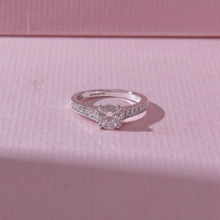 Top view of a white gold diamond engagement ring, featuring a round solitaire center stone with pavé-set shoulders. Captured against a soft pink backdrop, the design reflects timeless elegance and brilliance.
