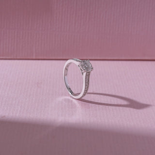 Angled view of a white gold diamond engagement ring, featuring a round solitaire center stone with pavé-set band detailing. Captured against a soft pink backdrop, the design reflects refined sparkle and elegance.
