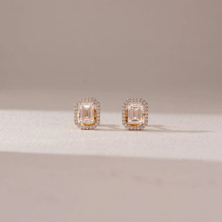 Front view of diamond stud earrings in 14KT yellow gold, featuring emerald-cut center diamonds with octagonal pavé halo settings, displayed on a beige surface highlighting symmetry and brilliance.
