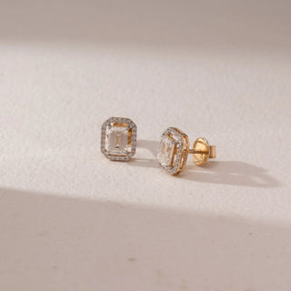 Angled view of diamond stud earrings in 14KT yellow gold, featuring emerald-cut center diamonds with octagonal pavé halo settings and screw backs, displayed on a beige surface highlighting detail and brilliance.
