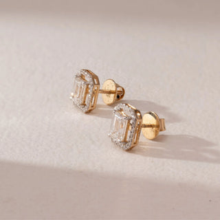 Side view of diamond stud earrings in 14KT yellow gold, featuring emerald-cut center diamonds with octagonal pavé halo settings and screw backs, set on a beige surface highlighting depth and sparkle.
