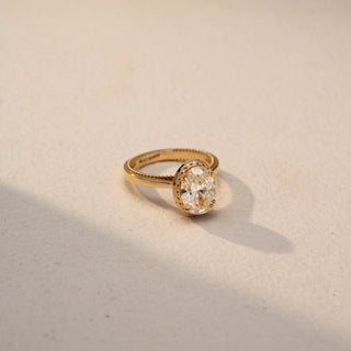 Side view of an oval solitaire ring in 14 KT Yellow Gold, featuring an oval-cut lab-grown diamond in a bezel-inspired setting with a delicate milgrain band, displayed against a beige backdrop.