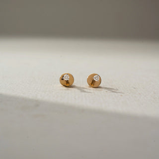 Front view of minimalist gold diamond stud earrings featuring tiny round diamonds set on polished circular gold discs, displayed on a soft neutral surface with subtle shadows.