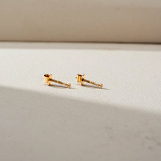 Side view of minimalist gold diamond stud earrings with tiny round diamonds in delicate prong settings, showcasing threaded screw-back posts on a soft neutral background.