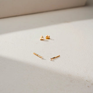 Angled view of minimalist gold diamond stud earrings with tiny round diamonds and threaded screw-back posts, displayed on a soft neutral surface with the backs placed in the background.