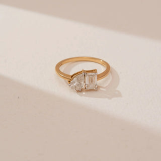 Angled top view of Reia’s 18 KT Yellow Gold Modern Twin Diamond Proposal Ring. Captured against a cream backdrop, the layout highlights the harmony of the emerald and pear-cut diamonds.