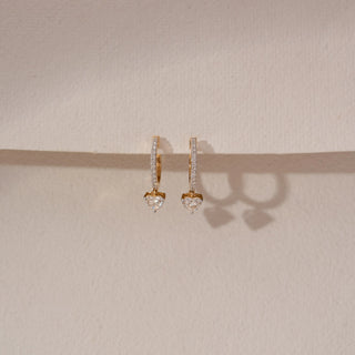 Front view of diamond hoop drop earrings in 14KT yellow gold, featuring pavé-set hoops with dangling heart-shaped diamonds, displayed against a beige backdrop highlighting delicate sparkle and elegance.

