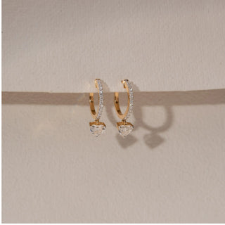 Side view of diamond hoop drop earrings in 14KT yellow gold, featuring pavé-set hoops with dangling heart-shaped diamonds, displayed against a beige backdrop highlighting depth and sparkle.
