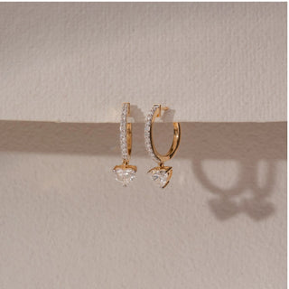 Angled view of diamond hoop drop earrings in 14KT yellow gold, featuring pavé-set hoops with dangling heart-shaped diamonds, displayed against a beige backdrop highlighting detail and brilliance.

