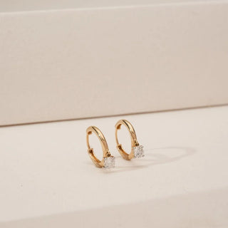 Angled view of yellow gold solitaire diamond hoop earrings featuring round lab-grown diamonds suspended at the base of sleek polished hoops on a neutral background.