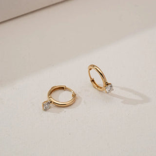 Angled view of yellow gold solitaire diamond hoop earrings with round lab-grown diamonds suspended at the base of smooth polished hoops, placed on a soft neutral studio surface.