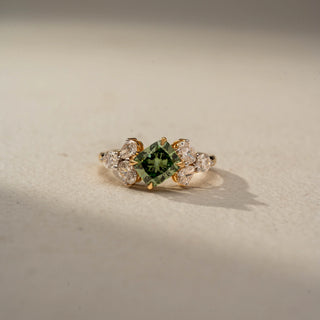 Front view of a gemstone engagement ring in 14 KT Yellow Gold, featuring a princess-cut green center stone flanked by pear-shaped lab-grown diamonds in a floral-inspired cluster setting, displayed against a beige backdrop.