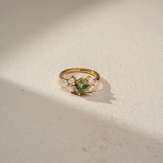 Top view of a gemstone engagement ring in 14 KT Yellow Gold, featuring a princess-cut green center stone flanked by pear-shaped lab-grown diamonds in a floral-inspired cluster setting, displayed against a beige backdrop.