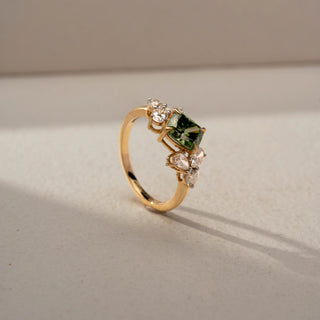Angled view of a gemstone engagement ring in 14 KT Yellow Gold, featuring a princess-cut green center stone flanked by pear-shaped lab-grown diamonds in a floral-inspired cluster setting, displayed against a beige backdrop.