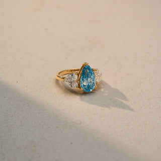 Side view of a three-stone ring in 14 KT Yellow Gold, featuring a pear-shaped blue center stone flanked by round lab-grown diamonds, displayed against a beige backdrop.