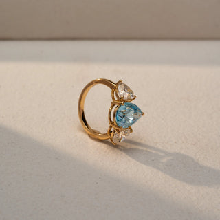 Angled view of a three-stone ring in 14 KT Yellow Gold, featuring a pear-shaped blue center stone flanked by round lab-grown diamonds, displayed against a beige backdrop.