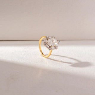 Angled view of Geometric Diamond Statement Ring in 14KT Yellow Gold, featuring a central diamond framed by baguette and round accents. Set on a beige surface, the design reflects bold structure and brilliance.

