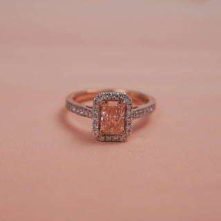 Front view of Reia Rose Halo Radiant Diamond Ring on a soft blush-peach background, showcasing a radiant-cut blush pink lab-grown diamond surrounded by a halo of white diamonds in 14KT rose gold.