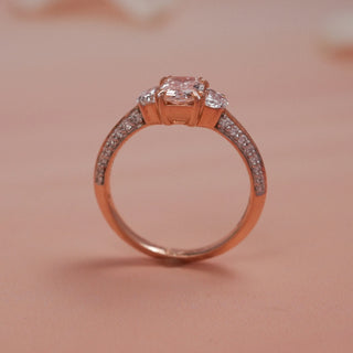 Side view of Reia Rose Glow Emerald Diamond Ring on a blush-peach background, highlighting the raised emerald-cut centre stone with half-moon sides and diamond-studded band in rose gold.