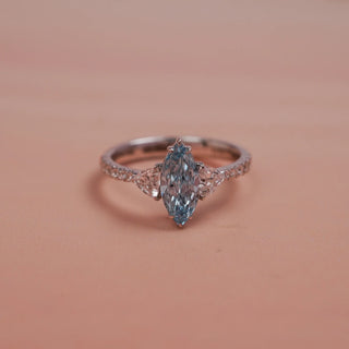 Side view of Reia Aqua Trine Marquise Diamond Ring on a soft blush-peach background, highlighting the elevated marquise-cut blue diamond centre stone with trilliant side accents set in 14KT white gold.