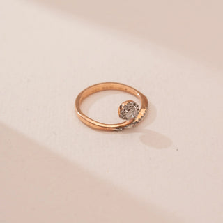 Top view of Reia’s 14 KT Rose Gold Graceful Swirl Diamond Ring. The centred round solitaire and its pavé-set swirling curve are showcased against a soft pink surface.