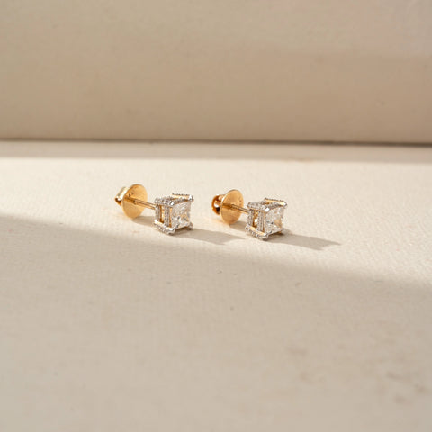 Side view of princess-cut diamond stud earrings in yellow gold, featuring square diamonds in elevated basket settings. Displayed on a beige backdrop, the profile showcases intricate craftsmanship.