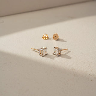 Angled view of princess-cut diamond stud earrings in yellow gold, featuring square diamonds in elevated basket settings with halo-inspired detailing. Displayed on a beige backdrop, the design sparkles elegantly.