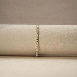 Front view of a diamond tennis bracelet in 14 KT Yellow Gold, featuring a continuous row of round brilliant diamonds in a sleek linear setting, displayed against a soft beige backdrop.
