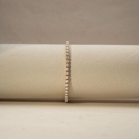 Front view of a diamond tennis bracelet in 14 KT Yellow Gold, featuring a continuous row of round brilliant diamonds in a sleek linear setting, displayed against a soft beige backdrop.