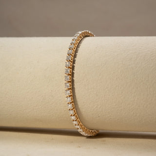 Close Upview of a diamond tennis bracelet in 14 KT Yellow Gold, featuring a continuous row of round brilliant diamonds in a sleek linear setting, displayed against a soft beige backdrop.